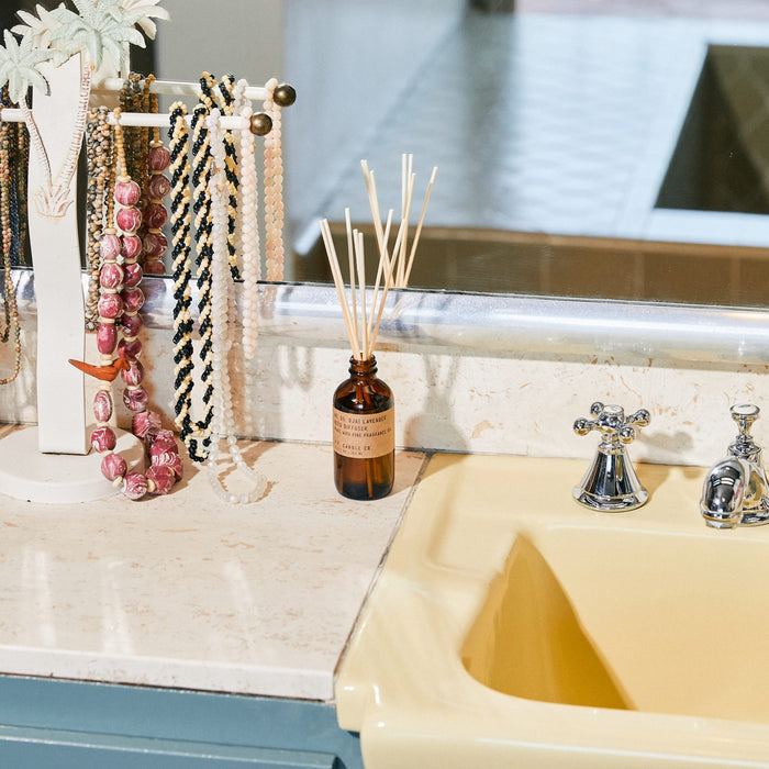 A bathroom countertop features a yellow sink, a vintage-style faucet, and a necklace rack. A P.F. Candle Co. Reed Diffuser in a brown bottle is placed beside the sink, while a large mirror reflecting tiles adorns the background.