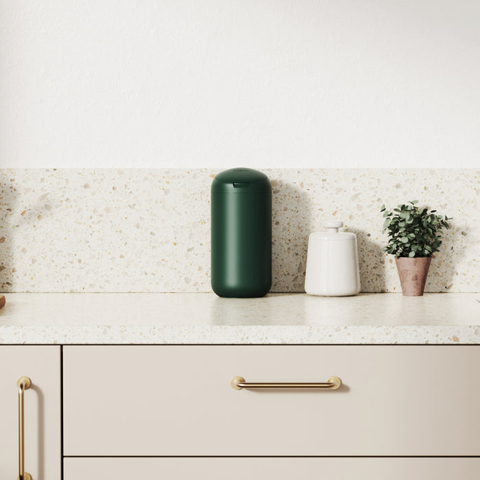 A minimalist kitchen counter displays a green cylindrical container, an airtight white jar with a lid, and a small potted plant. The light speckled surface pairs well with beige cabinets featuring gold handles, highlighted by Bioms award-winning Refillable Wipes Dispenser for style and practicality.