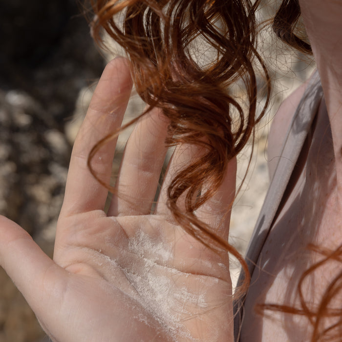 A close-up showcases a hand dusted with Refresh Dry Shampoo by Captain Blankenship. A lock of curly red hair drapes over the fingers, emphasizing its volume-boosting brilliance. The blurred background ensures the detailed foreground stands out.