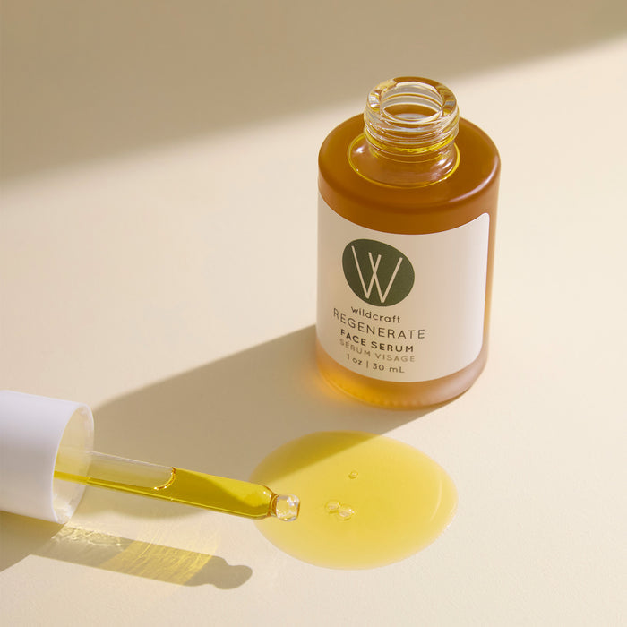 A bottle of Wildcraft Regenerate Face Serum, an anti-aging serum, rests on a light surface with an open dropper and pooled serum, softly lit by natural light.