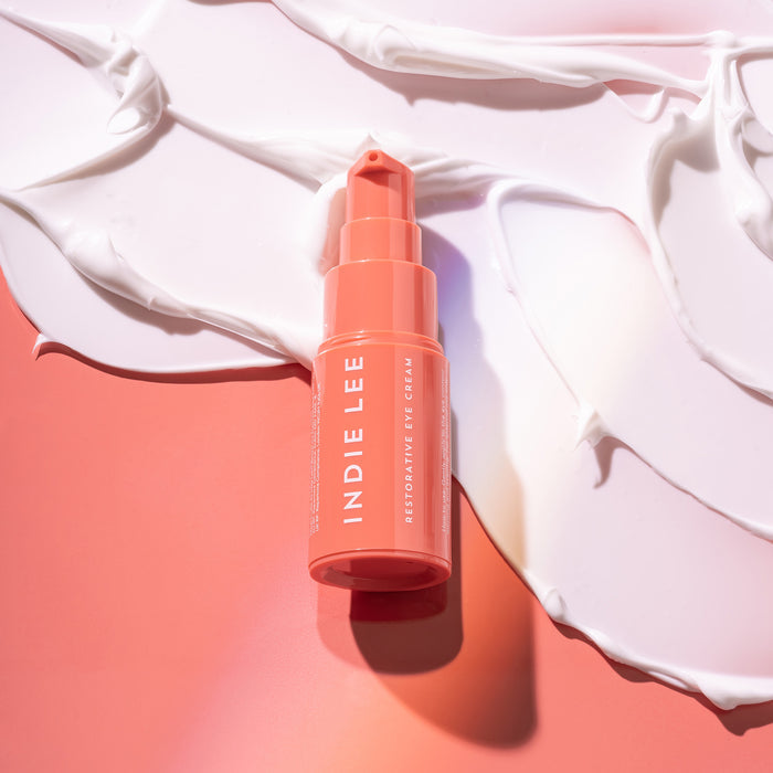A coral Indie Lee Restorative Eye Cream bottle, enhanced with Paracress extract, sits gracefully against swirled white cream on a coral background, creating a chic and vibrant contrast.