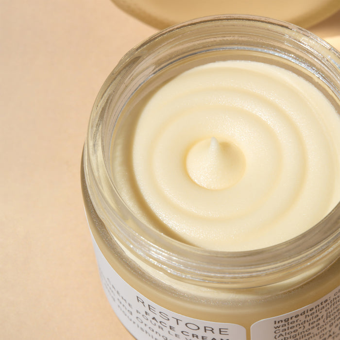 Open jar of the creamy, light yellow Restore Face Cream by Wildcraft, featuring a smooth, swirled texture ideal for dry skin. The RESTORE label softly shimmers through on a warm beige background, highlighting this handcrafted skincare marvel.