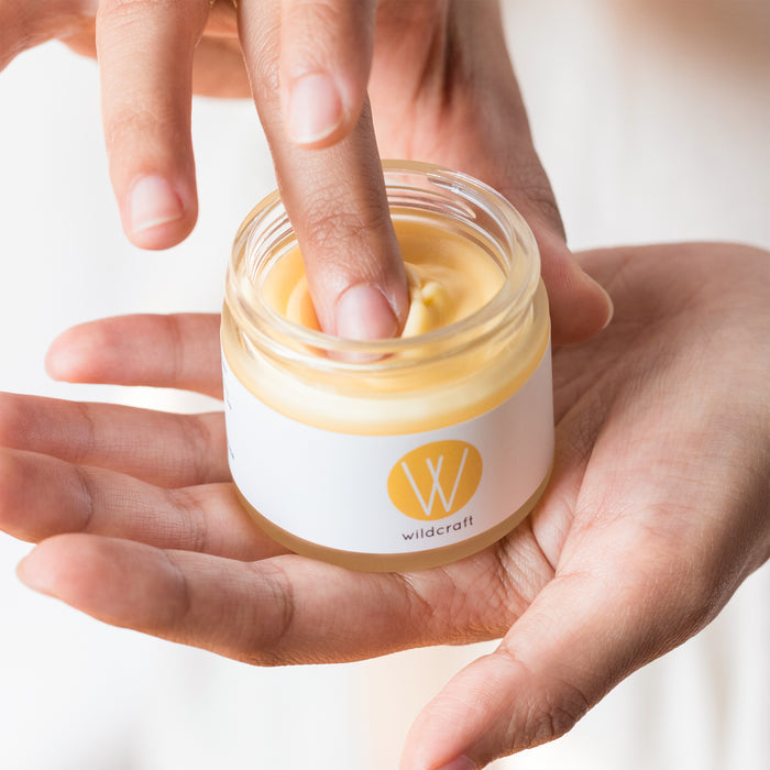A person holds a jar of Wildcrafts Restore Face Cream for dry skin, dipping a finger into its light yellow moisturizer. The jar, adorned with the Wildcraft logo, is showcased in soft lighting, highlighting this handcrafted skincare moment.