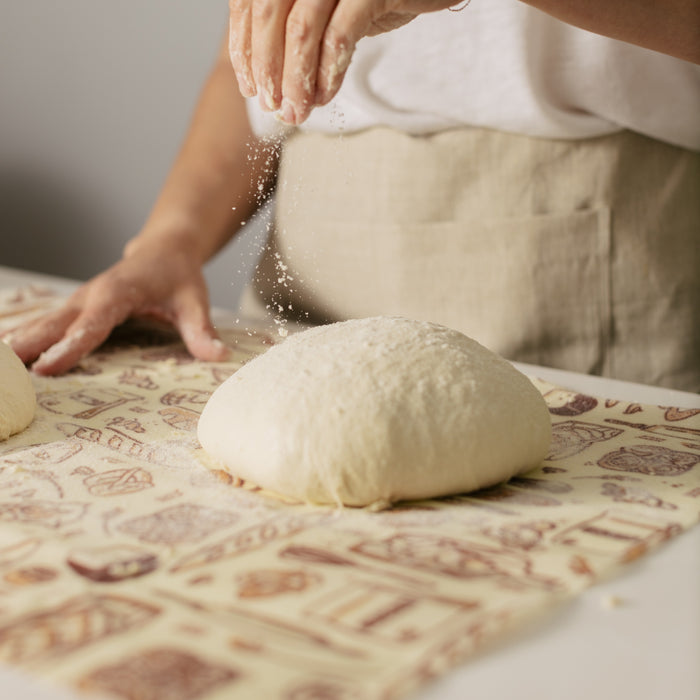 A person in a white shirt and beige apron sprinkles flour over sourdough on a patterned cloth, prepping it for baking. Nearby, a Bees Wrap Bread Wrap helps reduce single-use kitchen waste.