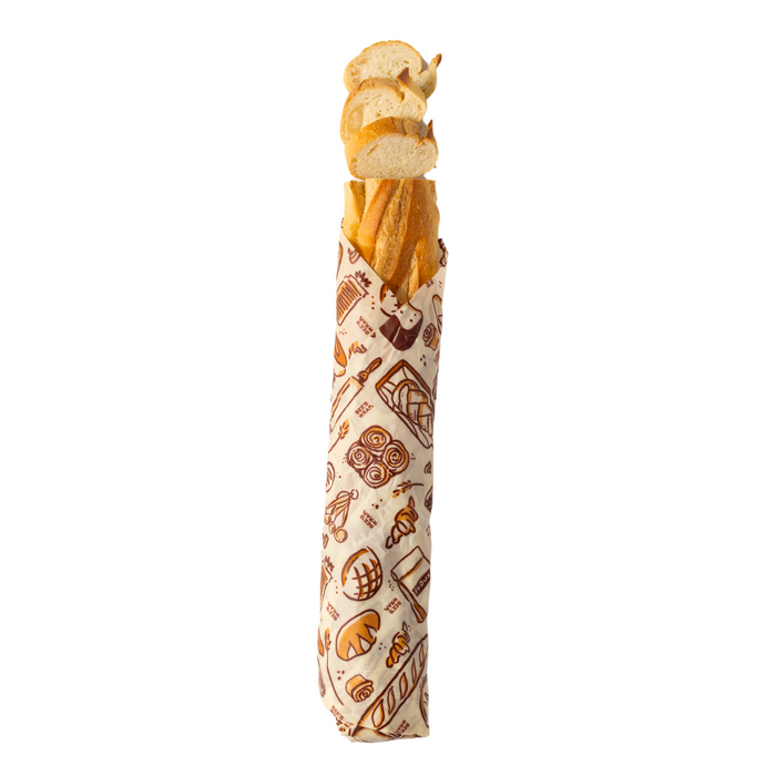 A Bees Wrap Bread Wrap decorated with a bread-themed pattern wraps a baguette, with several sourdough baguette slices on top, all displayed against a white background.