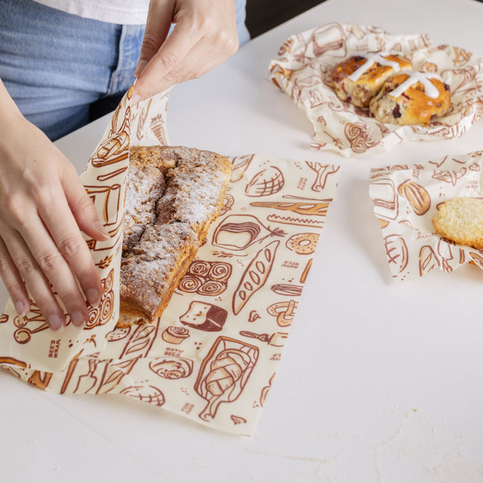 Someone wraps a fresh loaf in a Bees Wrap Bread Wrap on a white surface. Other baked goods wrapped in Bees Wrap to help reduce waste are arranged nearby.