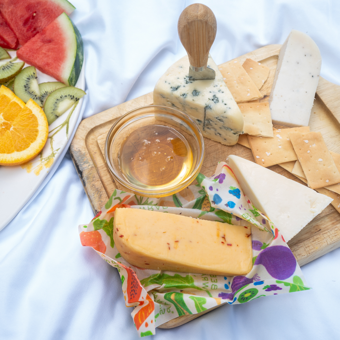 A wooden board with assorted cheeses, crackers, and honey sits on a white cloth beside a plate of fresh fruit, all kept fresh with Bees Wrap Assorted Wraps-5 pack.