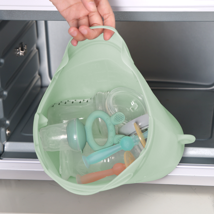 A hand places a light green Haakaa Reusable, Silicone Multipurpose Steamer & Steriliser Bag containing baby bottles, nipples, and rattles into a dishwasher. The dark gray interior is open, showcasing the essentials neatly arranged for easy microwave sterilization.