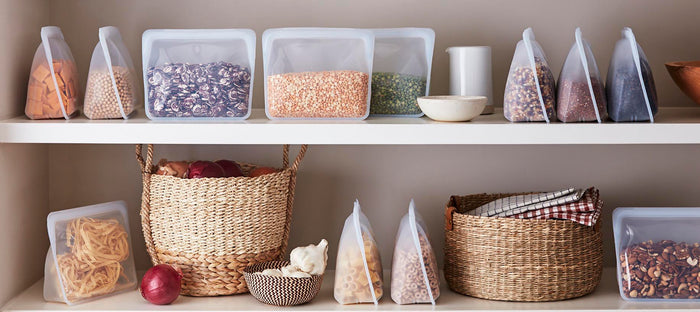 Two shelves display eco-friendly products: the top holds Stasher Reusable Silicone Stand-Up Bags with grains and beans, while the bottom has onions, a garlic bulb, pasta in a bag, and a woven basket with cloth. Neutral tones create an organized look.