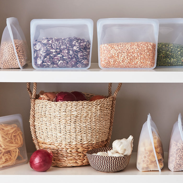 An eco-friendly pantry is organized using Stashers Reusable Silicone Stand-Up Bags, displaying dried chickpeas, dark beans, lentils, and pasta. A woven basket holds red onions and a small bowl contains garlic bulbs for efficient organization.