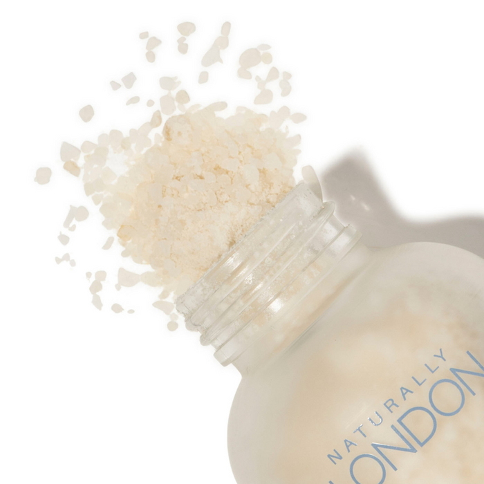 A glass bottle of Naturally Londons Revitalizing Foot Soak spills coarse white salts onto a white surface—ideal for moisturizing foot soaks and relieving dry, itchy skin.