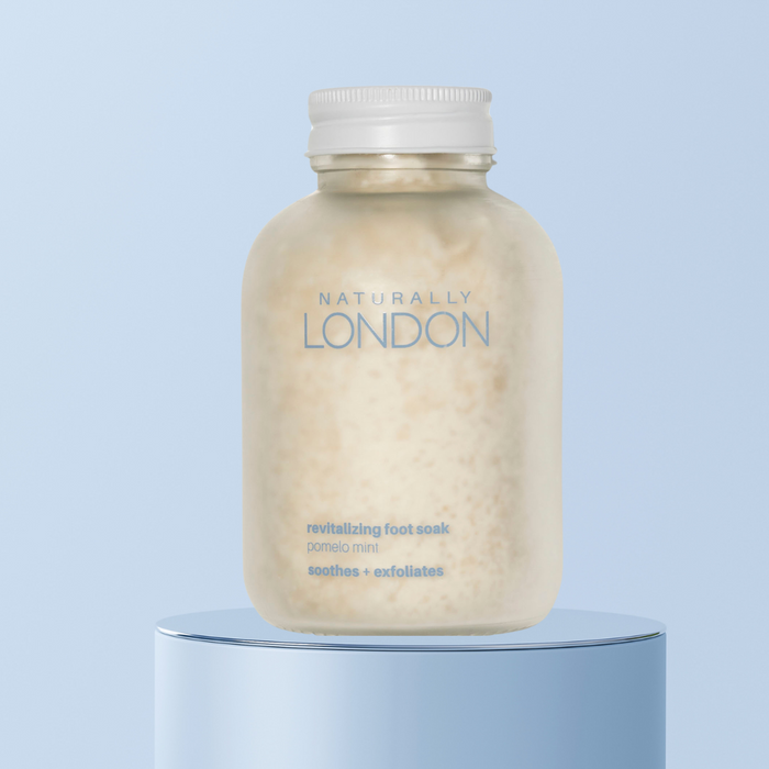 A jar of Naturally London Revitalizing Foot Soak sits on a light blue reflective pedestal against a blue background, revealing its pale granular contents.