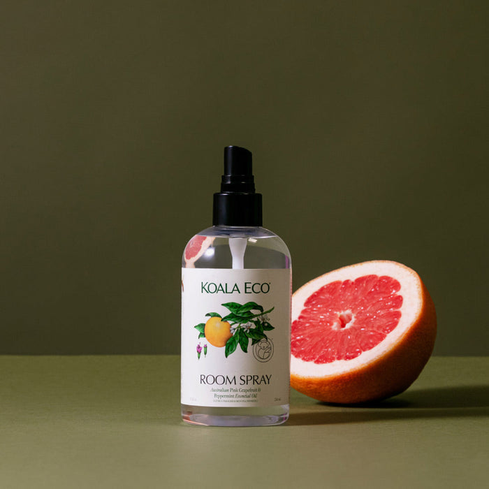 A Koala Eco Room Spray bottle, featuring a citrus fruit illustration on its label, stands beside a halved grapefruit against an olive-green background, evoking the invigorating scent of the Australian bush.
