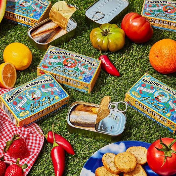 Fishwifes Sardines with Preserved Lemon lie on grass, encircled by colorful produce - tomatoes, lemons, an orange, strawberries, red peppers - and crackers on a checkered cloth. The tin showcases bright retro designs.