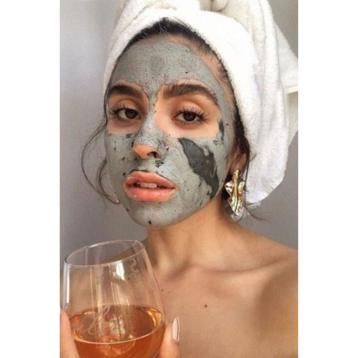 A woman, relaxed in a wrapped towel and gold earrings, enjoys a self-care moment with the Sea-el Sea Kelp Mud Mask on her face while holding a glass of light-colored wine.