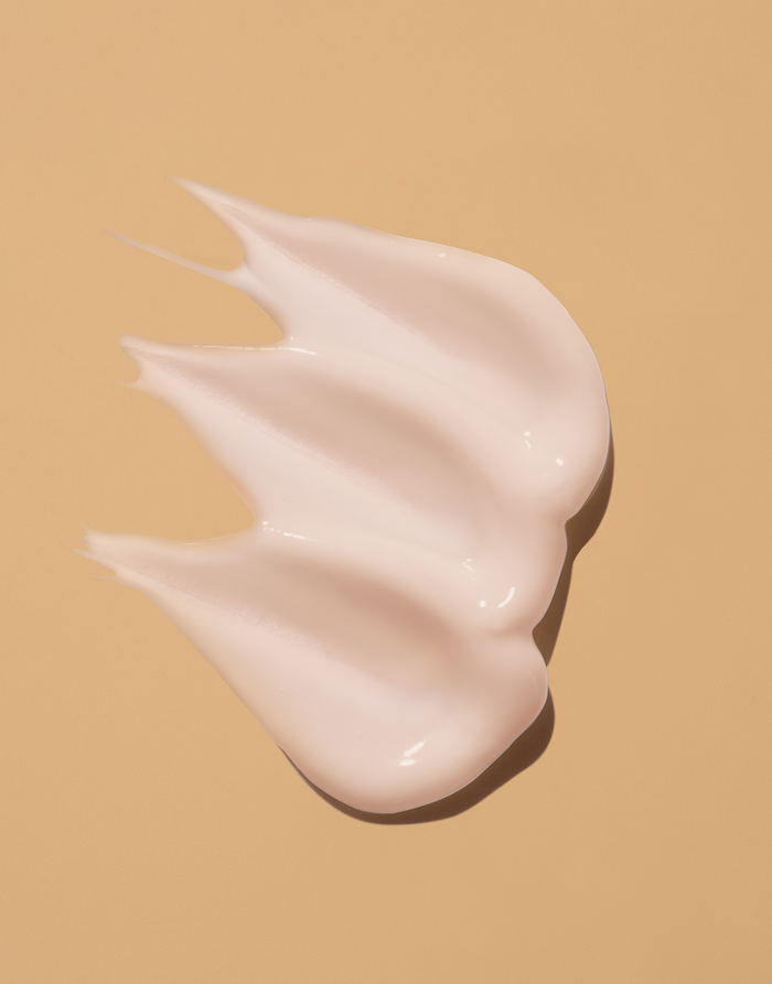 A swirl of Seaglow Overnight Serum by OSEA, a creamy AHA-infused lotion in light pink, is artistically spread on a beige backdrop, creating a smooth texture with three distinct peaks.