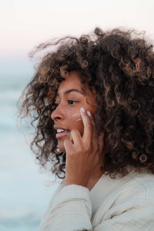 With curly hair, a person applies OSEAs Seaglow Overnight Serum to their cheek, enjoying an AHA treatment for smoother skin while standing by the ocean. Theyre clad in a light-colored jacket against a blurred horizon and pastel sky backdrop.