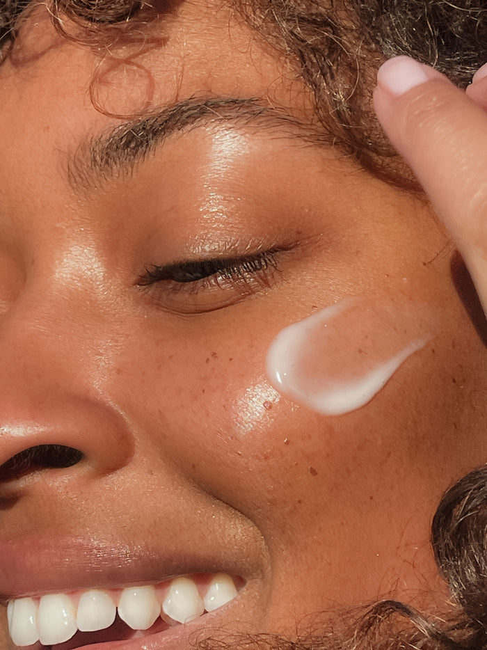 Close-up of a person smiling with their eyes closed, applying OSEAs Seaglow Overnight Serum on their cheek. Their skin glows, hinting at a smoother complexion, and their fingernail is painted light pink.