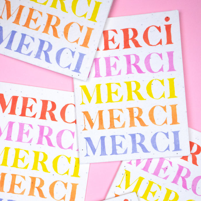 On a pink background, Seed Cards by Supra Endura showcase the word MERCI in bold colors like red, pink, yellow, orange, and purple. These playful cards are crafted from plantable greeting cards that blossom into something beautiful.