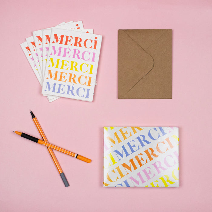 A flat lay on a pink surface displays colorful Supra Endura Seed Cards with a stack of plantable greeting cards, a brown envelope, two orange pens, and a gift wrapped in the same eco-friendly design, adding sustainability to your correspondence.