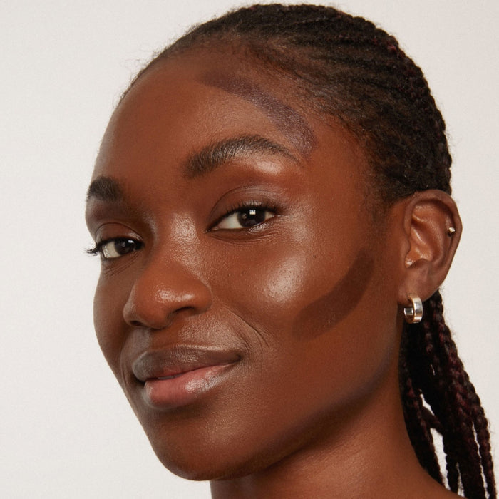 A person with braided hair and small hoop earrings looks forward, smiling slightly. Axiologys Shape Sticks are expertly used to apply contour makeup on their cheeks, forehead, and above the brow against a plain white background.
