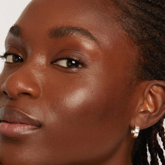 Close-up of a person with smooth, glowing skin, enhanced by Axiologys Shape Stick, wearing a small silver hoop earring. The plain background emphasizes the face, showcasing natural beauty and serenity.