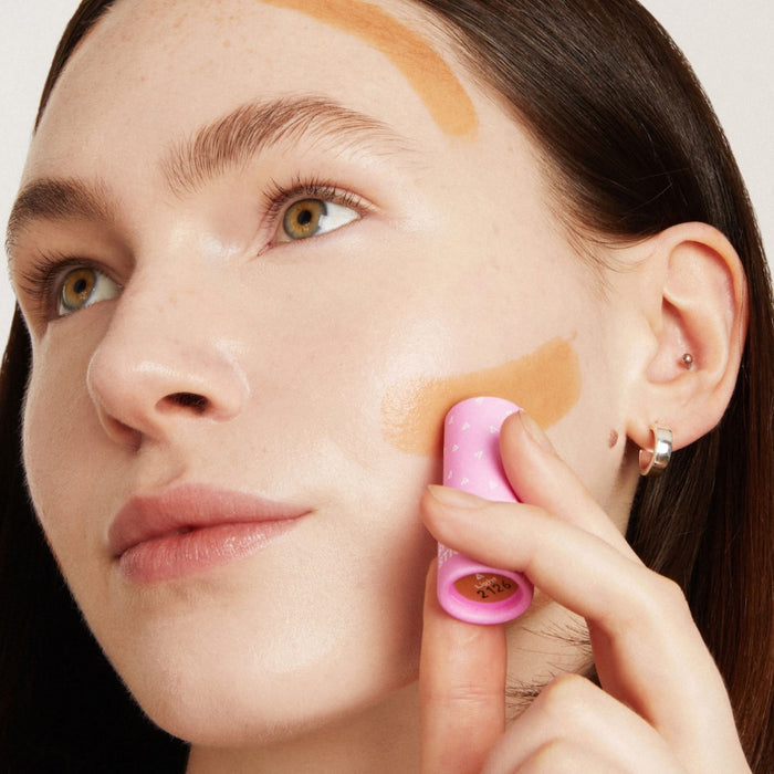 Someone with fair skin applies Axiologys Shape Stick to their cheek. The pink stick, featuring a small heart pattern, provides broad swipes of hydrating long-wear foundation. They have brown hair and light brown eyes.