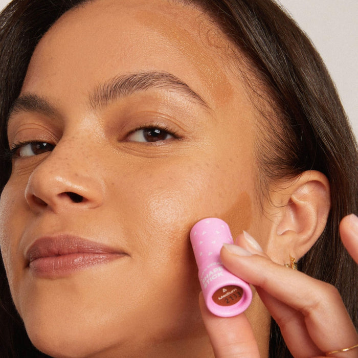 A person applies Axiologys Shape Stick for long-wear hydrating coverage to their cheek, revealing its light brown hue from the pink tube. With long dark hair and a slight smile at the camera, they stand against a plain, light background.
