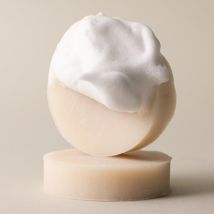 A round, beige Leaf Shave Shave Soap bar with creamy lather sits atop another identical bar, both set against a simple, light background.