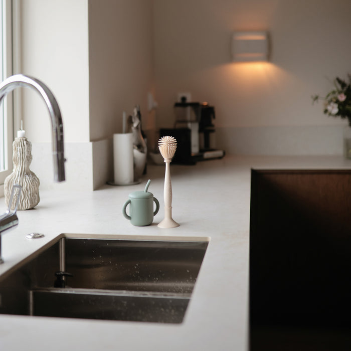 A minimal kitchen counter features a sink, cup with straw, mushie Silicone Baby Bottle Brush – Dual-Head, Non-Scratch, BPA-Free, candles, coffee machine, and flower vase. Warm lighting enhances the cozy atmosphere.