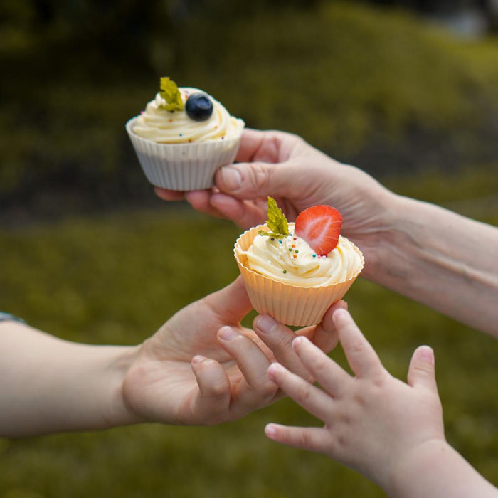 Two hands present cupcakes in vibrant Net Zero Co. Silicone Baking Cups, one garnished with a blueberry and mint and the other with a strawberry and mint. A childs hand reaches eagerly for these tempting treats against a blurred outdoor background.