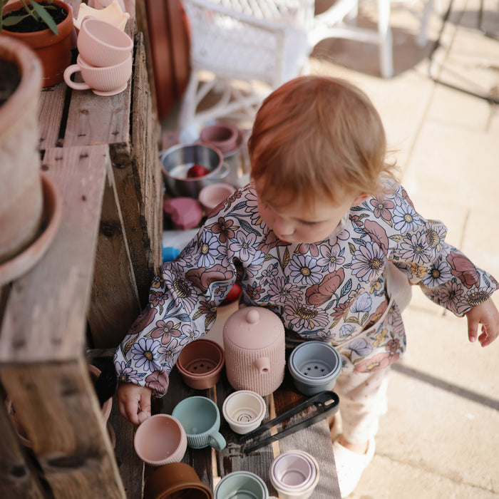 A young child in a floral shirt enjoys pretend play with the mushie Silicone Tea Play Set – Pretend Play, BPA-Free, featuring pastel-colored silicone cups and teapot on a wooden shelf outdoors, surrounded by potted plants.