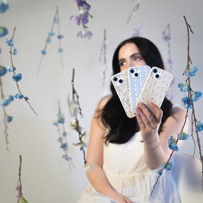 A woman in a white dress displays three Single Case Activation Codes by Pela Case, each featuring customizable floral designs. Blue and purple floral patterns create a whimsical, eco-friendly vibe with a softly blurred background.