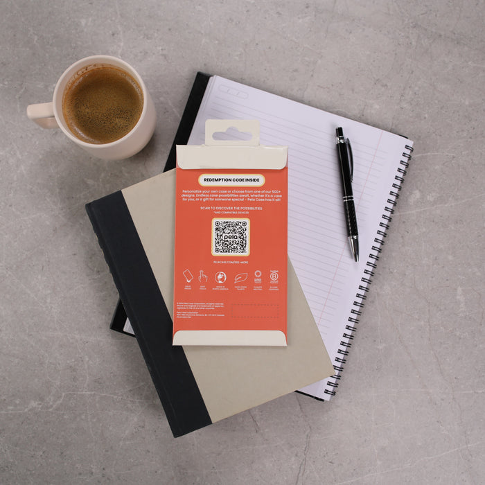A coffee cup sits beside a customizable gray notebook with a black pen on top, an open spiral notebook, and a Pela Case Single Case Activation Code package featuring a red QR code section. This elegant setup rests on the gray marble surface, lending an eco-friendly touch to your workspace.