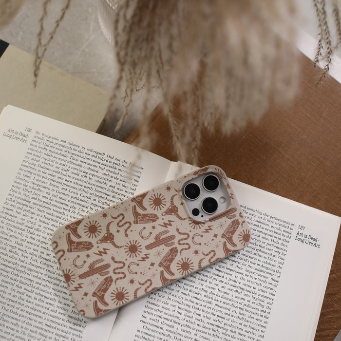 A Pela Case, featuring an eco-friendly celestial pattern, protects a smartphone resting on an open book. The setting is cozy and neutral-toned, with soft brown surfaces and blurred dried flowers enhancing the backdrop.