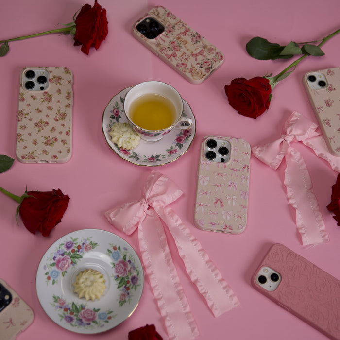 Aesthetic scene featuring Pela Cases floral and customizable pink iPhone cases made from sustainable materials, complemented by red roses, a yellow-filled teacup, a white flower-shaped treat, and pink bows on a soft pink background.