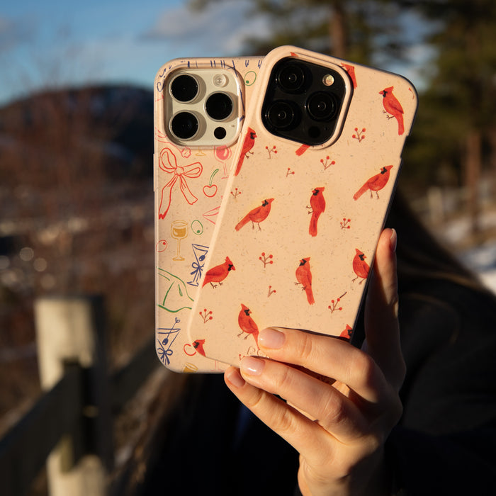 A person outdoors holds two smartphones, one with a customizable beige Pela Case featuring red cardinals and the other with a cream Pela Case adorned with colorful doodles made from sustainable materials, against a backdrop of trees, a fence, and mountains under a clear blue sky.