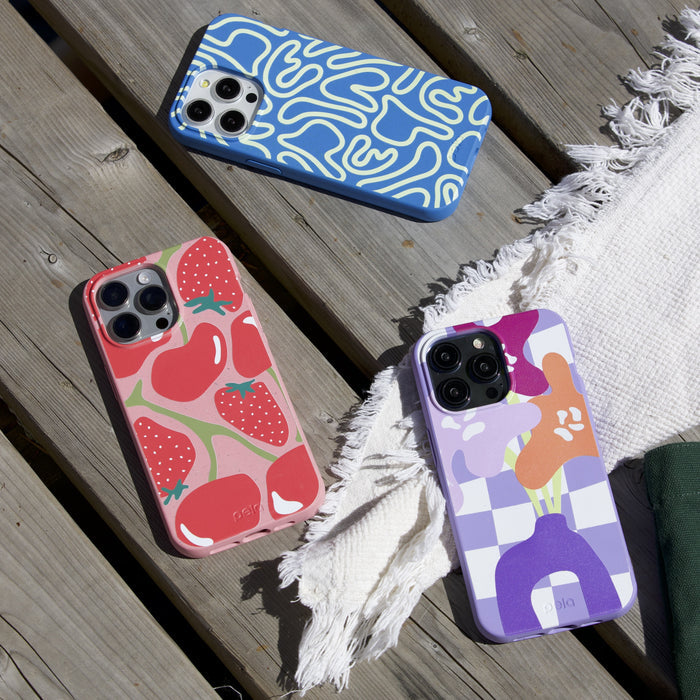 Three Pela smartphones featuring eco-friendly, customizable cases rest on a wooden surface next to fringed white fabric. The cases display abstract blue designs, red strawberries, and multicolored floral checkered patterns.