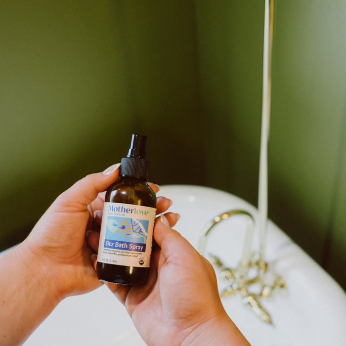 A person holds a bottle of Motherlove Sitz Bath Spray above a white tub with gold fixtures and a green wall, showcasing its soothing postpartum recovery benefits.