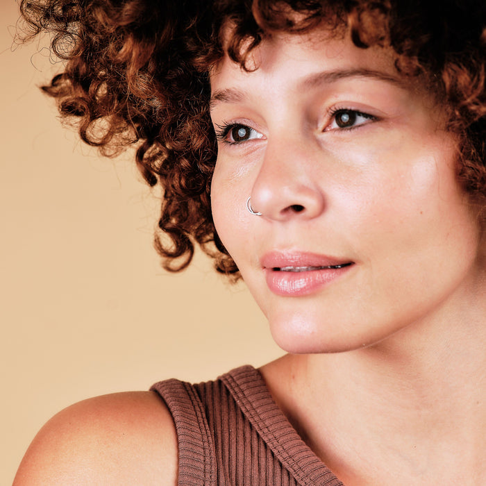 A person with curly hair and a nose ring gazes sideways, calm in expression. They wear a brown sleeveless top that highlights their Skin Recovery Serum-treated complexion by Thrive Natural Care, set against a soft beige background.