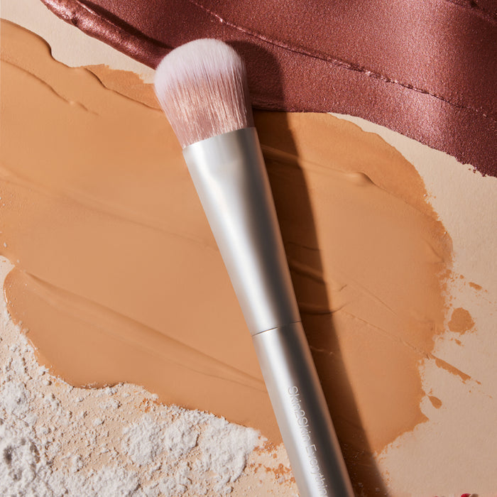A luxe RMS Beauty Skin2Skin Everything Brush with a silver handle rests on a surface covered in beige foundation swatches, white powder, and shimmery bronze fabric.