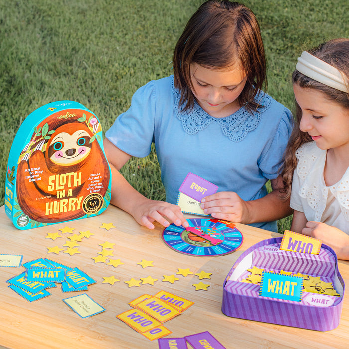 Two young girls play eeBoo Piece & Love’s Sloth in a Hurry Spinner Game at a wooden table, holding cards and a colorful spinner, with game pieces and boxes labeled WHO and WHAT encouraging creativity and imagination.