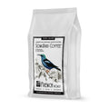 A white Songbird Coffee bag by Thanksgiving Coffee shows a vibrant bird design and florals. The text reads Very Dark, French Roast, Smoky, Rich, Intense. This Bird Friendly Coffee features the American Birding Association logo—symbolizing sustainable excellence.