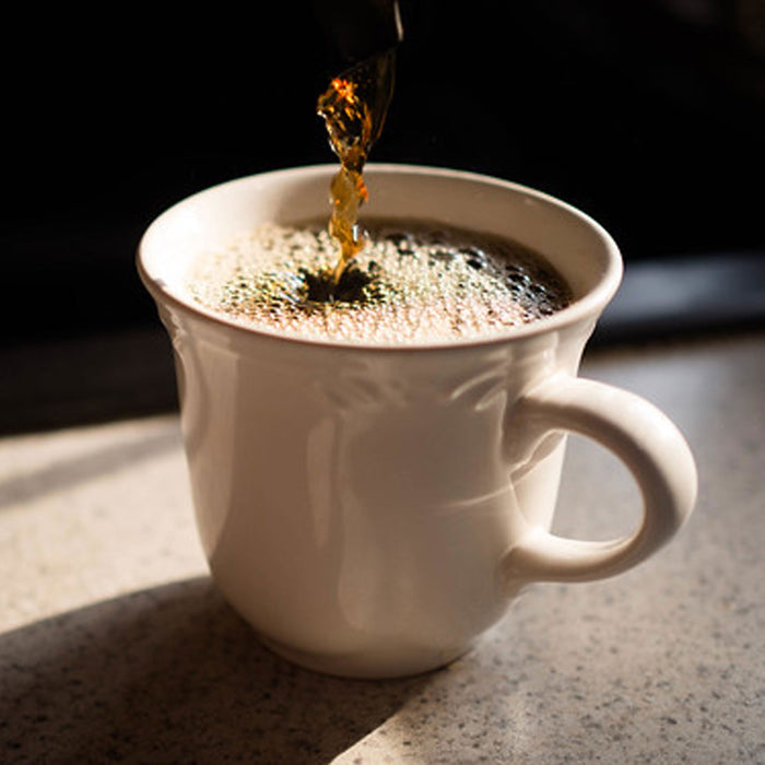 A white mug on a countertop is being filled with Thanksgiving Coffees Songbird French Very Dark, capturing light reflections on the bubbling surface.