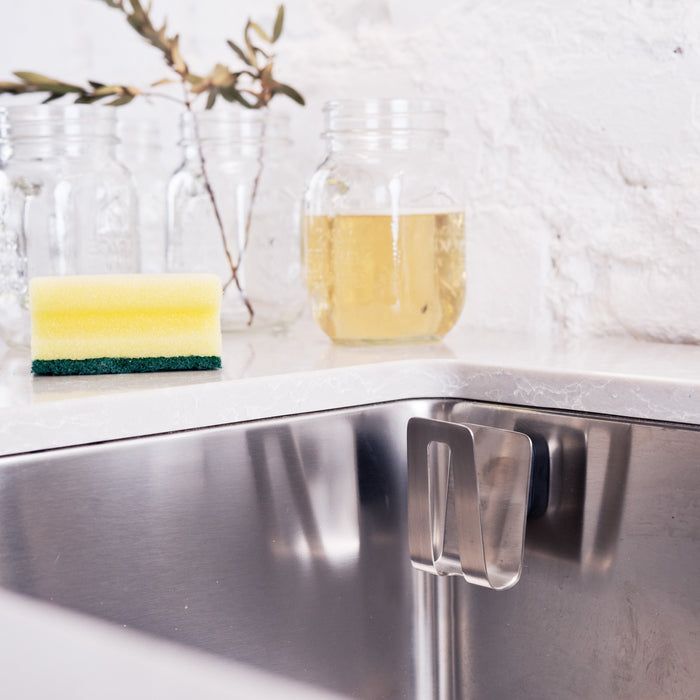 A stainless steel sink includes the HAPPY SiNKS Sponge Holder with a Magnet Fitting System for hygienic storage. A yellow-green sponge is on the counter, with glass jars and a jar of yellow liquid against a white brick wall in the background.