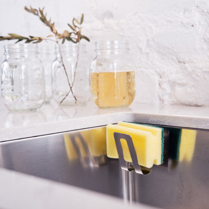 On the countertop, framed by a stark white brick wall, rests a HAPPY SiNKS Sponge Holder providing hygienic storage with an eye-catching yellow and green sponge. Nearby, three glass jars filled with water and twigs enhance the kitchens charm and functionality.