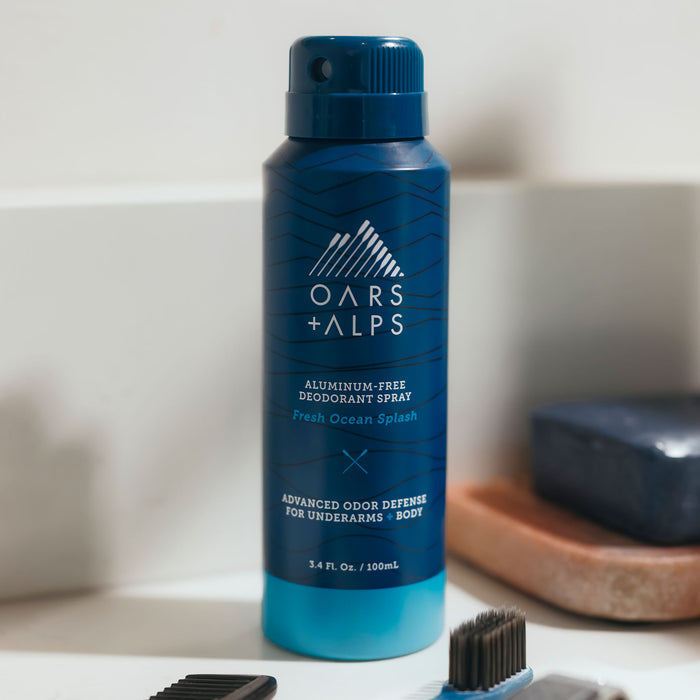 An Oars + Alps Spray Deodorant in a blue can is on a bathroom counter beside a hair clipper and bar of soap.