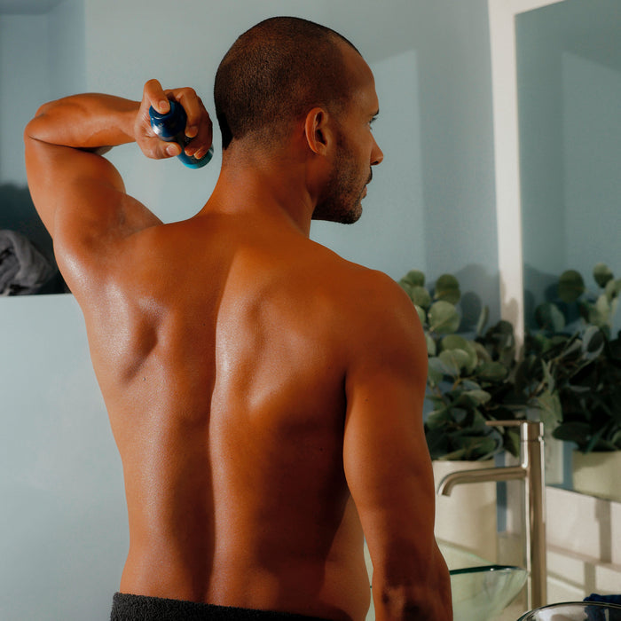 A man with a towel around his waist uses Oars + Alps Spray Deodorant in California Coast Scent in a bathroom with light blue walls and a plant on the counter, enjoying its refreshing aroma as he gets ready.