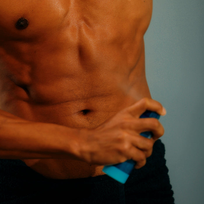 A person sprays Oars + Alps Spray Deodorant onto their bare torso while wearing black shorts, set against a plain background.