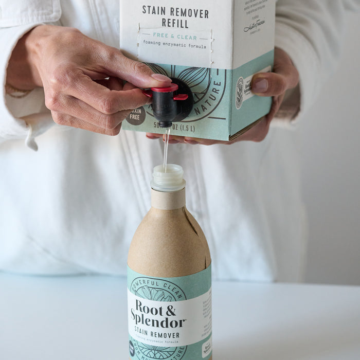 Wearing a light-colored shirt, a person pours Root and Splendor Stain Remover Refill into a reusable bottle labeled Root & Splendor, choosing this eco-friendly solution.